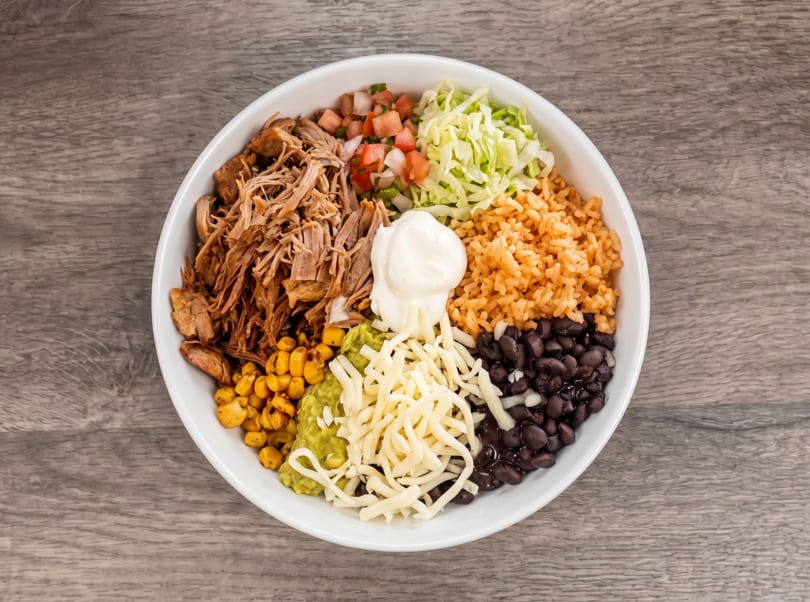 Overhead view of a burrito bowl with pulled pork, rice, beans, corn, guacamole, cheese, and sour cream.