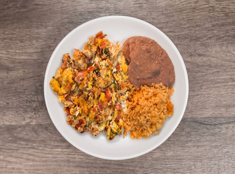 Scrambled eggs with tomatoes and onions, refried beans, and Spanish rice on a white plate.