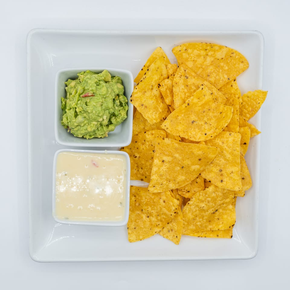 A white rectangular plate holds tortilla chips, a bowl of guacamole, and a bowl of queso dip.
