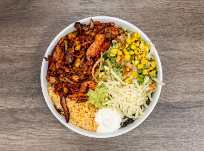 Overhead view of a colorful burrito bowl with rice, meat, corn, lettuce, guacamole, cheese, and sour cream.