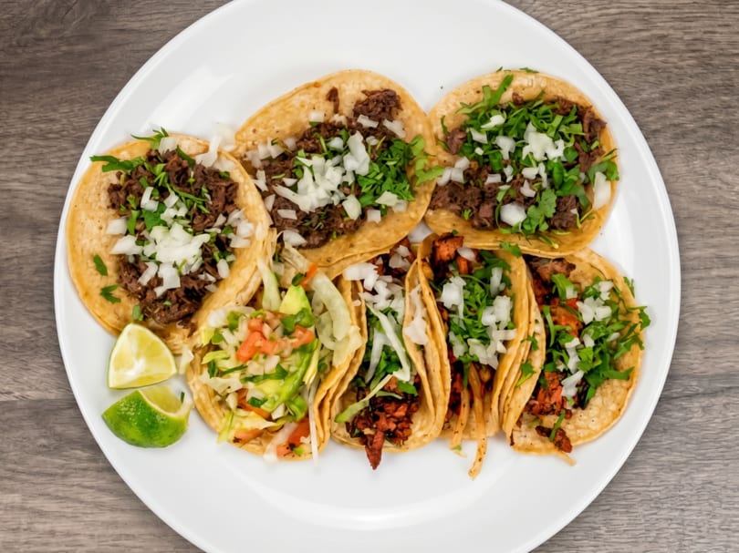 A white plate holds seven tacos with various fillings, cilantro, onions, and two lime wedges.