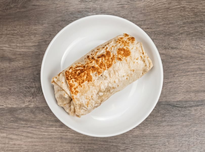 A grilled burrito with a lightly browned tortilla rests on a white plate on a wooden table.