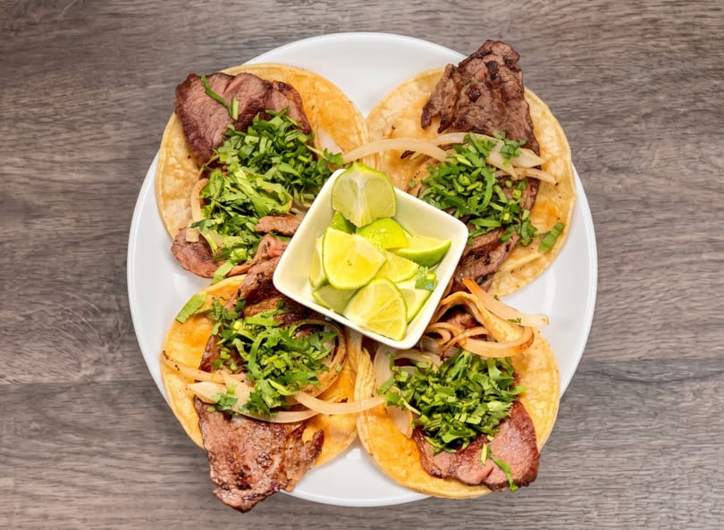 Four steak tacos with cilantro, onions, and lime wedges on a white plate.