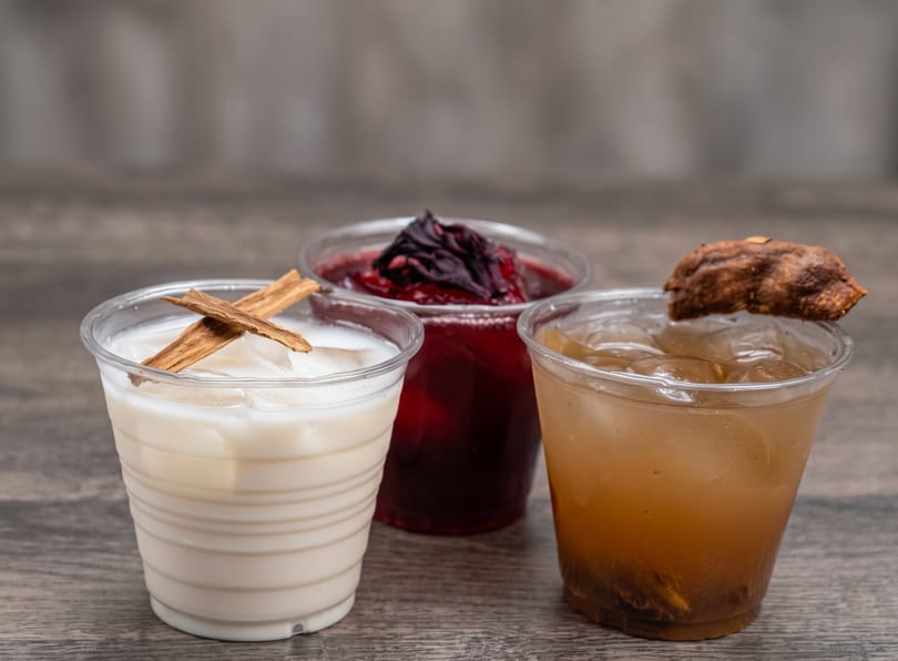 Three plastic cups of horchata, jamaica, and tamarindo drinks on a wooden surface.