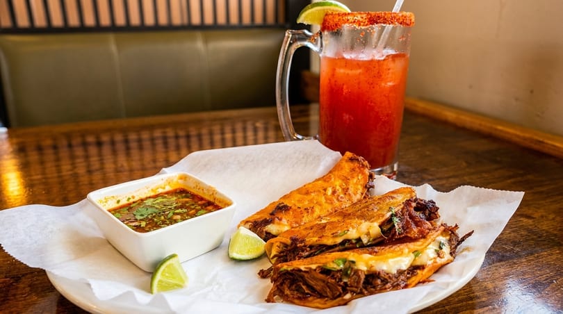 Three birria tacos with melted cheese, a side of consommé, and a michelada on a wooden table.