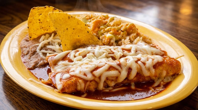 Steaming enchiladas with melted cheese, rice, refried beans, and tortilla chips on a yellow plate.